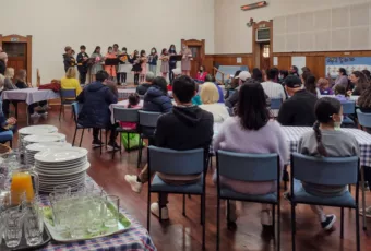 A lively community dinner at Puriri Centre Community Hub Takapuna, with people of diverse ages and ethnicities seated at long tables, enjoying meals and conversations.